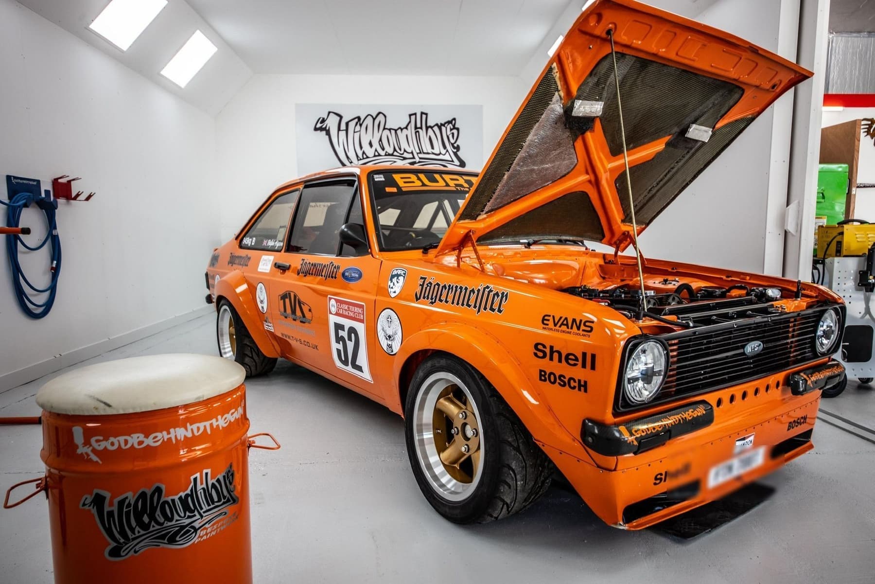 Orange Ford Escort Mk2 rally car in Jägermeister livery, bonnet raised, under the lights in the Willoughbys spray booth